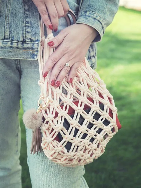 A close-up of a person's hand holding a stylish, oversized woven tote bag in a natural tone, standing in a brightly lit, minimalist entryway, with sunlight emphasizing the bag's texture and modern design.