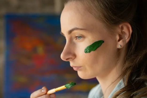A makeup artist's hand delicately applying a green color corrector stick to a model's cheek, targeting a small area of redness. The application is precise and minimal.